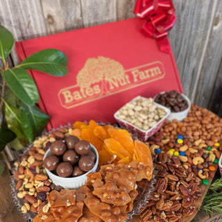 A gourmet nut and candy gift spread from Bates Nut Farm, featuring an assortment of pecans, almonds, peanuts, chocolate-covered nuts, dried apricots, caramel candy, and colorful candies arranged on a wooden board in front of a red gift box with a red bow.