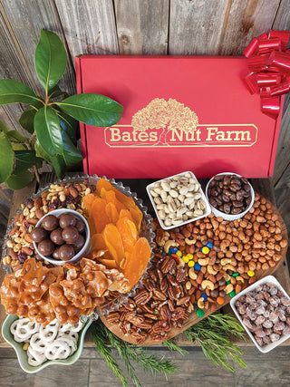 A gourmet nut and candy gift board from Bates Nut Farm, featuring a large red gift box with the brand logo, surrounded by assorted nuts, dried apricots, pretzels, chocolate-covered nuts, and candy on a rustic wooden background.